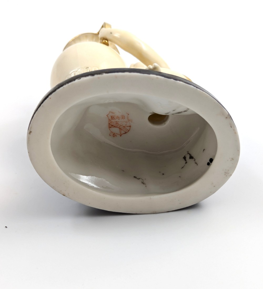 An ivory-coloured porcelain figurine depicting a small child seated beside a gilded amphora vessel on a dark base. View IX