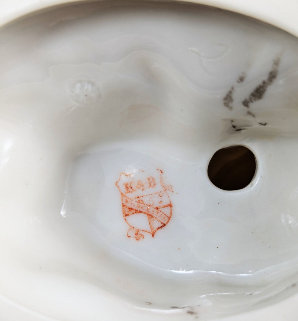 An ivory-coloured porcelain figurine depicting a small child seated beside a gilded amphora vessel on a dark base. View X