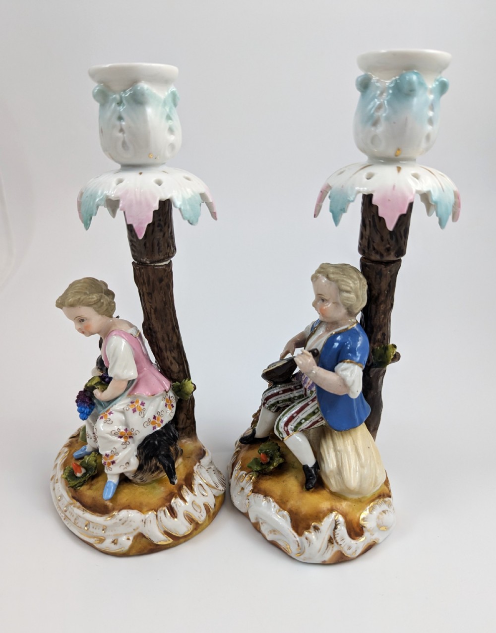 A pair of hand-painted German Sitzendorf porcelain figural candlesticks featuring a shepherdess and a lute player seated beside tree trunks supporting leafy sconces. View II