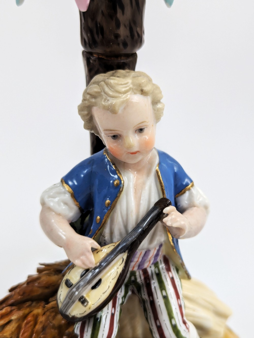 A pair of hand-painted German Sitzendorf porcelain figural candlesticks featuring a shepherdess and a lute player seated beside tree trunks supporting leafy sconces. View X