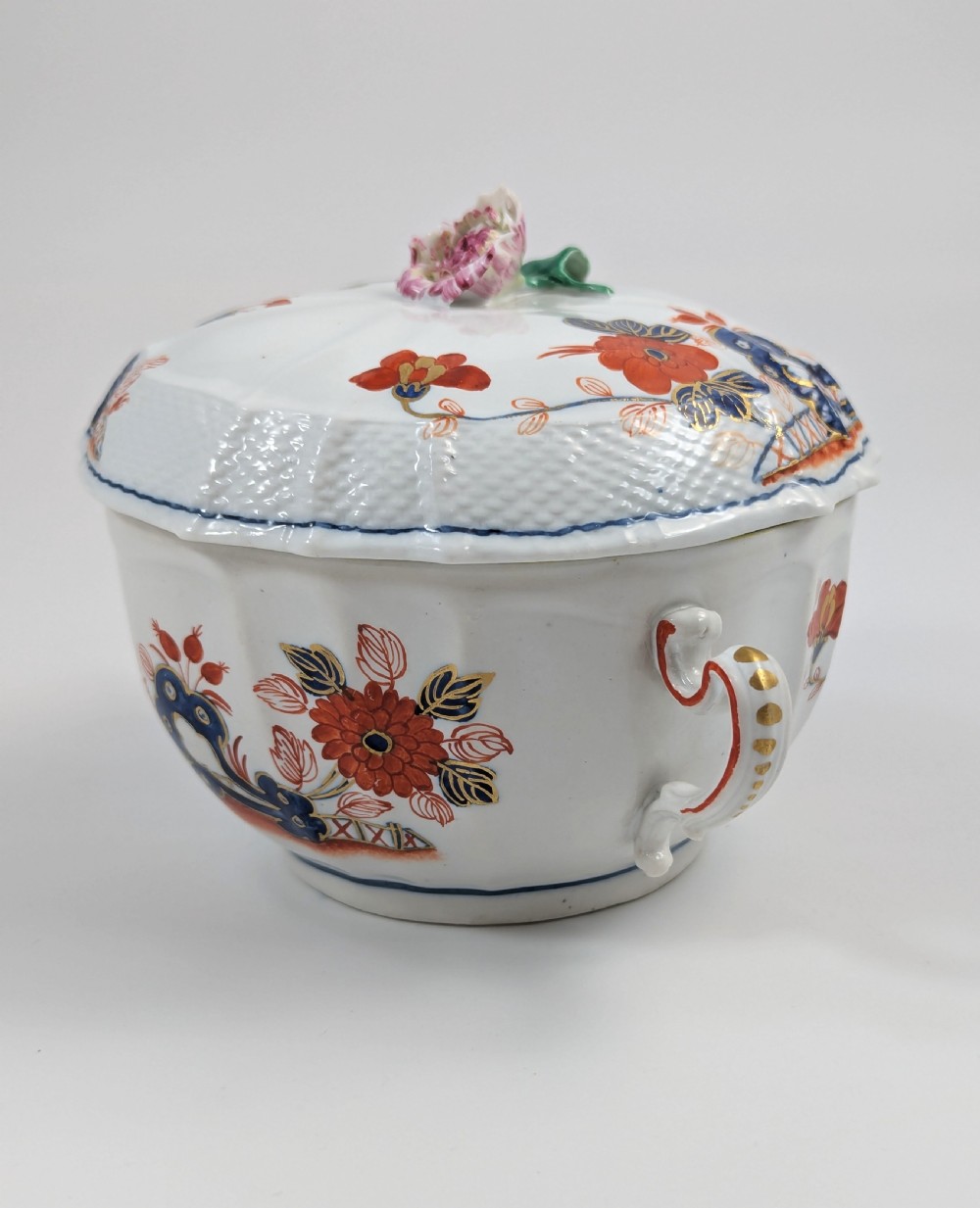 An antique white porcelain lidded broth cup decorated with hand-painted red and blue floral motifs, featuring twin scroll handles and a sculpted floral lid finial. View II