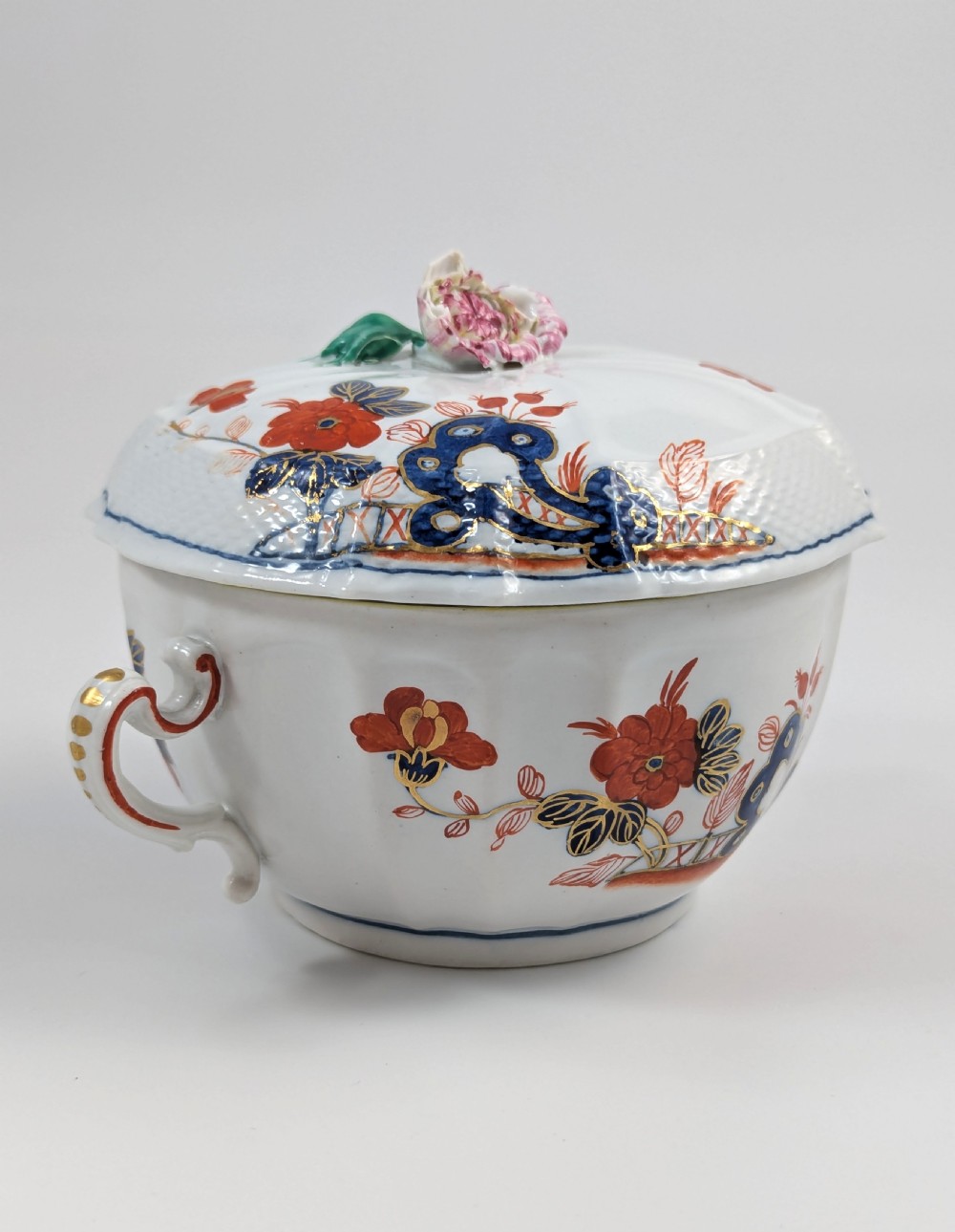 An antique white porcelain lidded broth cup decorated with hand-painted red and blue floral motifs, featuring twin scroll handles and a sculpted floral lid finial. View III