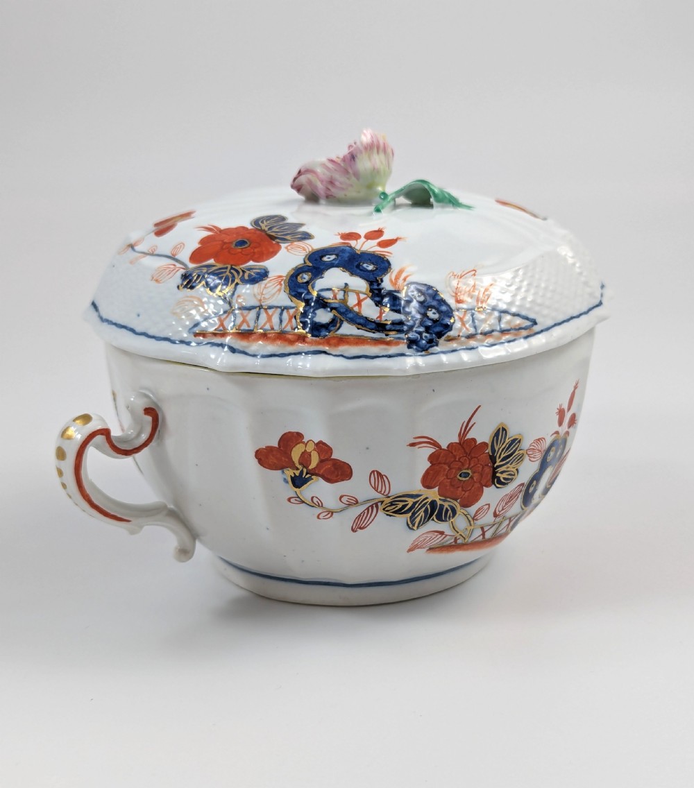 An antique white porcelain lidded broth cup decorated with hand-painted red and blue floral motifs, featuring twin scroll handles and a sculpted floral lid finial. View V