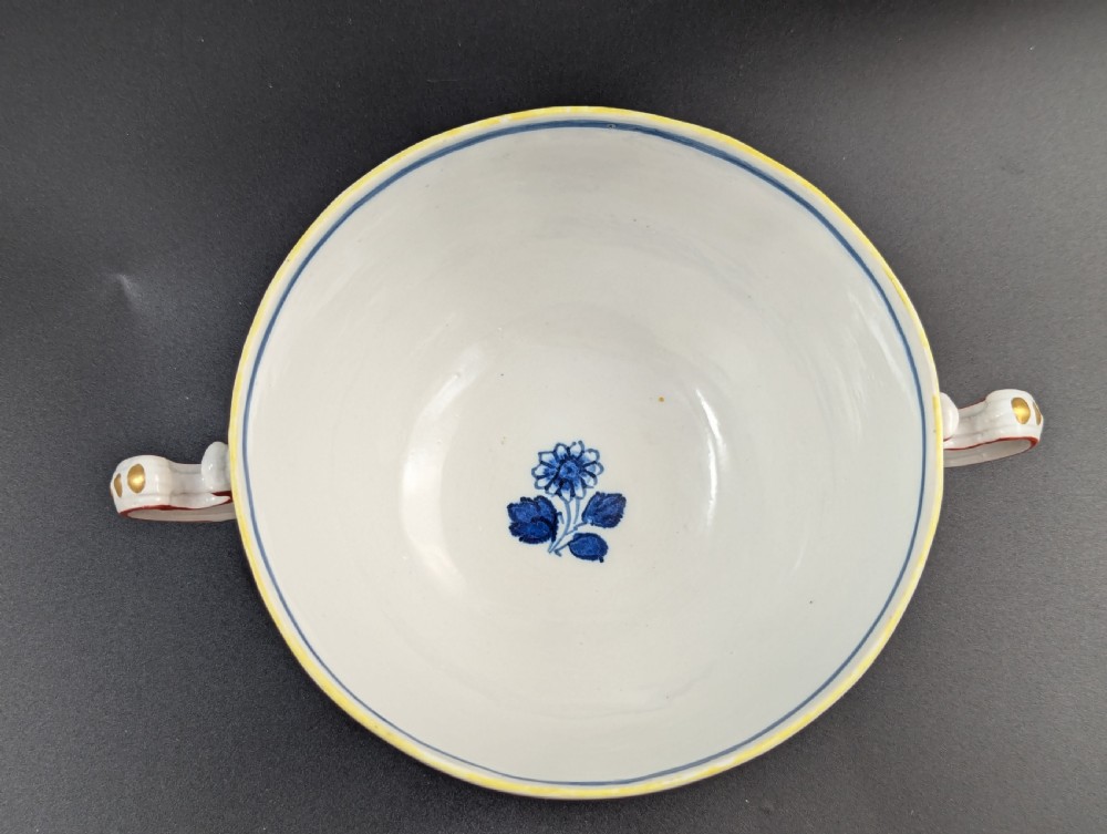 An antique white porcelain lidded broth cup decorated with hand-painted red and blue floral motifs, featuring twin scroll handles and a sculpted floral lid finial. View X