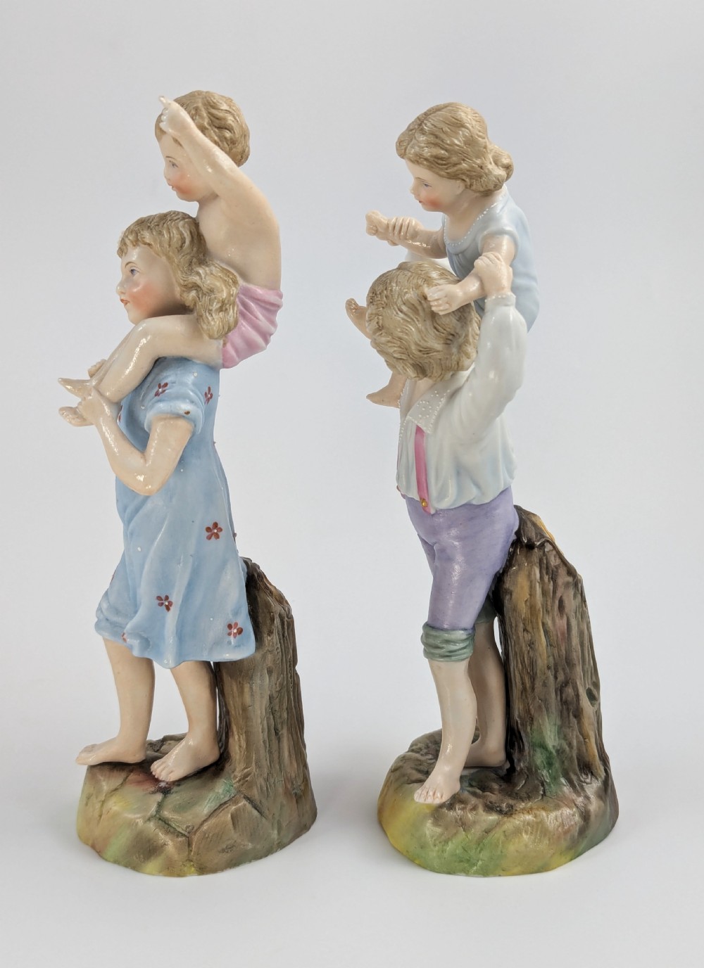 A pair of hand-painted German porcelain figurines depicting a boy and a girl carrying younger children piggyback style on tree stump bases. View II