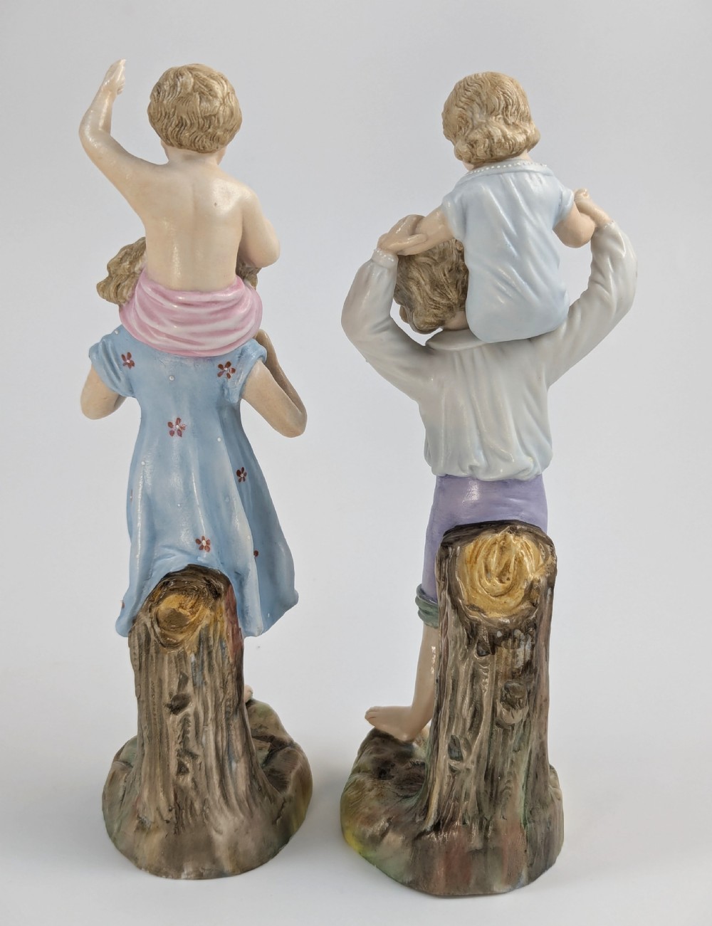 A pair of hand-painted German porcelain figurines depicting a boy and a girl carrying younger children piggyback style on tree stump bases. View III