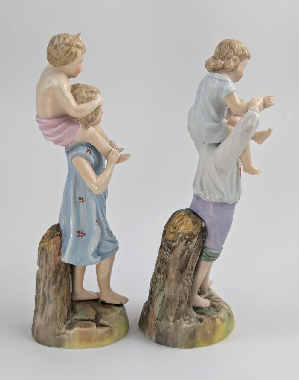 A pair of hand-painted German porcelain figurines depicting a boy and a girl carrying younger children piggyback style on tree stump bases. View IV
