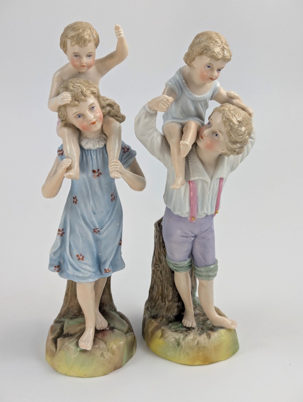 A pair of hand-painted German porcelain figurines depicting a boy and a girl carrying younger children piggyback style on tree stump bases. View V