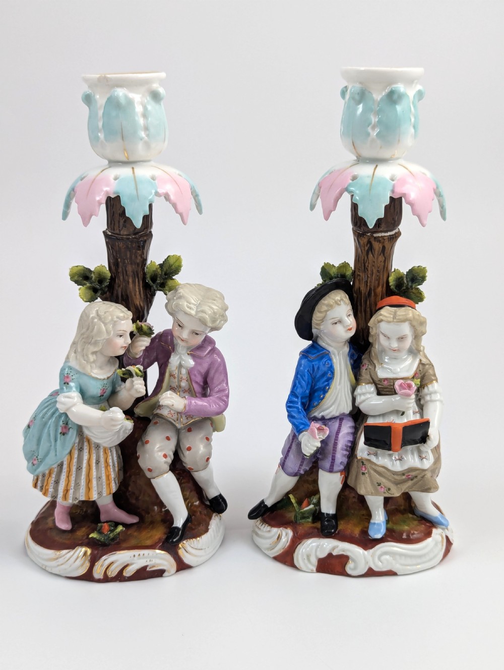 A pair of hand-painted porcelain figural candlesticks depicting eighteenth-century styled children beside floral tree columns.