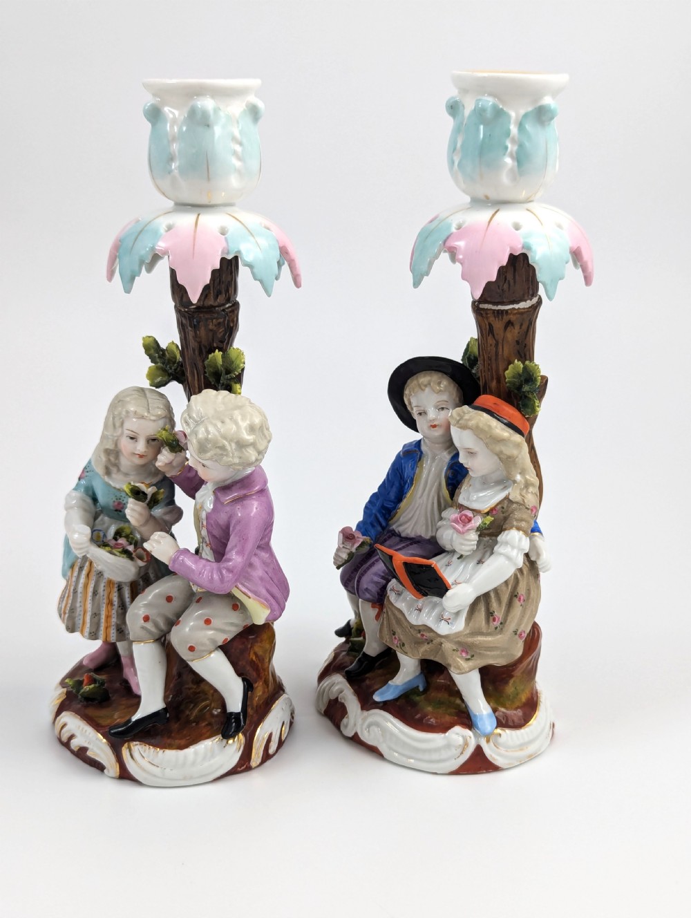A pair of hand-painted porcelain figural candlesticks depicting eighteenth-century styled children beside floral tree columns. View II