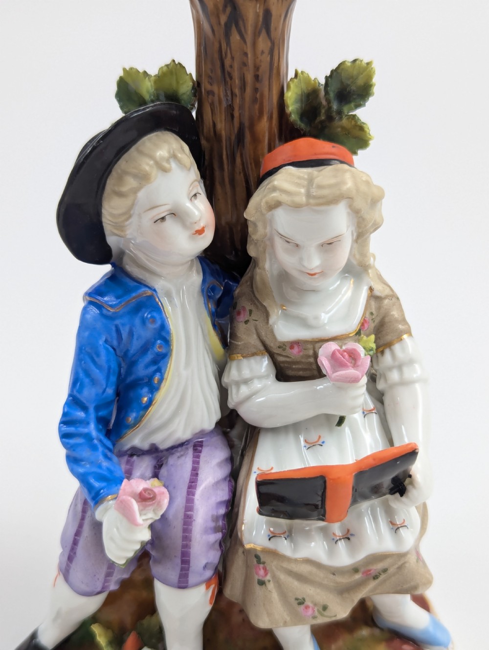 A pair of hand-painted porcelain figural candlesticks depicting eighteenth-century styled children beside floral tree columns. View IX