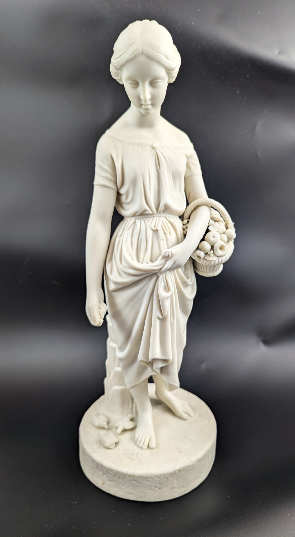An off-white Parian porcelain figurine depicting a standing classical female figure holding a fruit basket on a circular base.