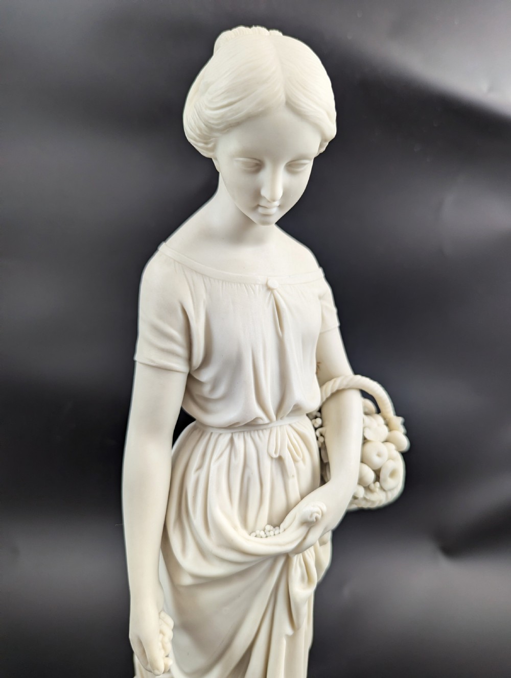 An off-white Parian porcelain figurine depicting a standing classical female figure holding a fruit basket on a circular base. View VIII