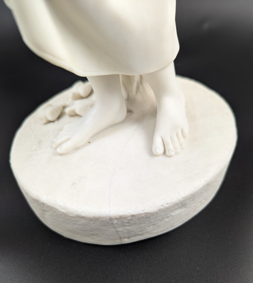 An off-white Parian porcelain figurine depicting a standing classical female figure holding a fruit basket on a circular base. View X