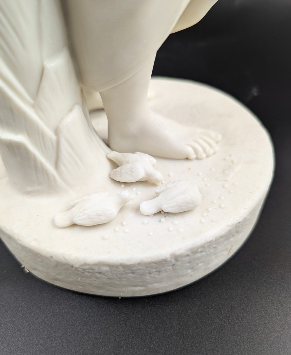 An off-white Parian porcelain figurine depicting a standing classical female figure holding a fruit basket on a circular base. View XI