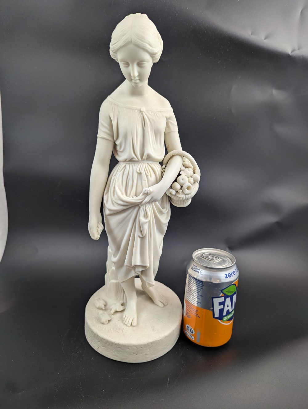 An off-white Parian porcelain figurine depicting a standing classical female figure holding a fruit basket on a circular base. View XIII