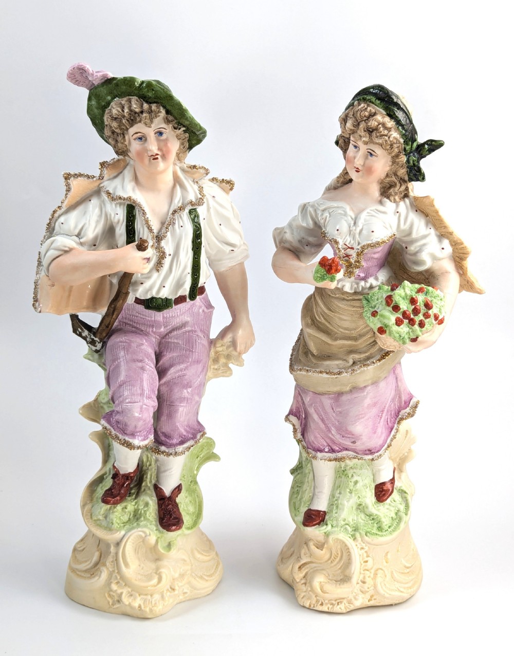 A pair of antique German bisque porcelain figurines depicting a man and woman in detailed pastoral attire with gilded accents on scroll bases.
