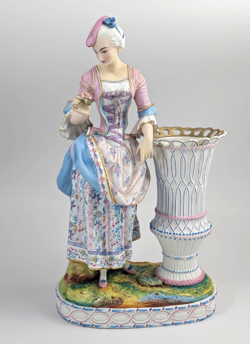 A 19th-century French bisque porcelain figural vase featuring a female figure in pastel Rococo costume standing beside a lattice-patterned vase on a painted base.