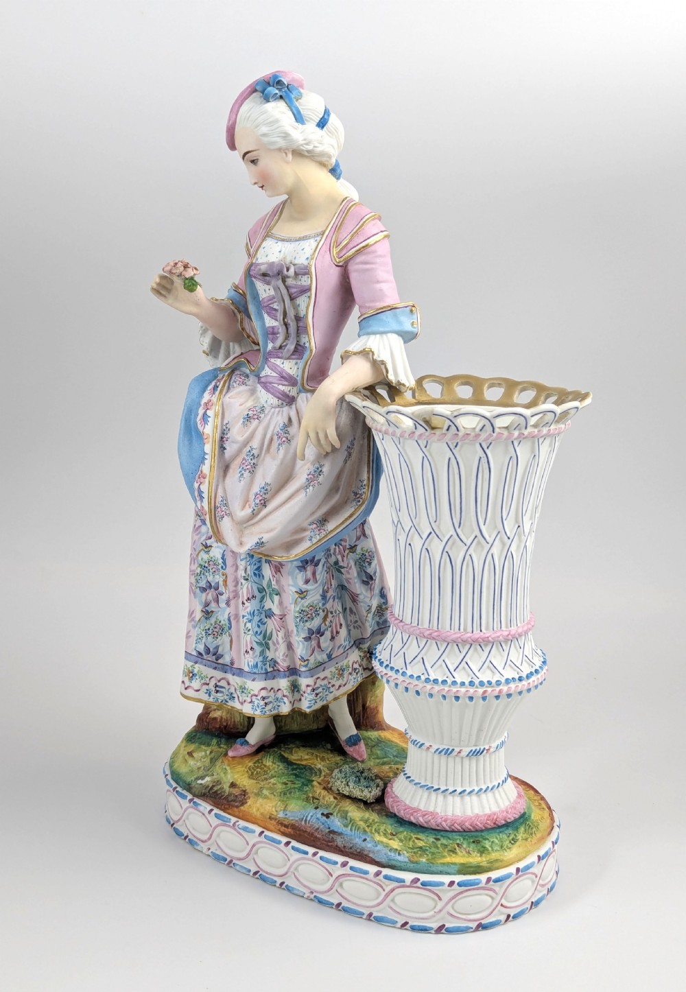A 19th-century French bisque porcelain figural vase featuring a female figure in pastel Rococo costume standing beside a lattice-patterned vase on a painted base. View II