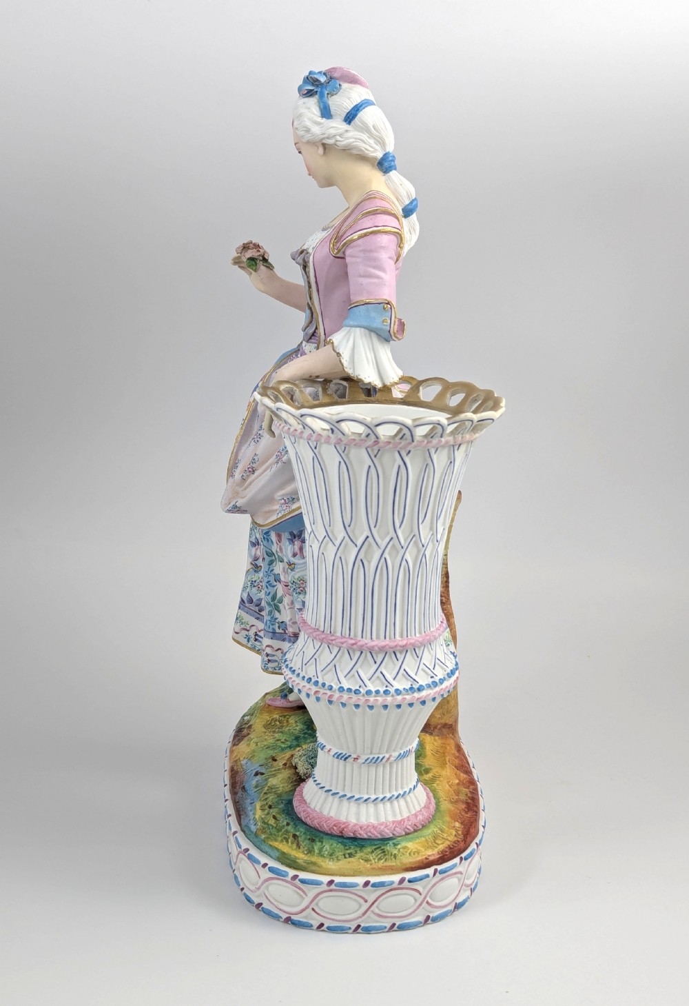 A 19th-century French bisque porcelain figural vase featuring a female figure in pastel Rococo costume standing beside a lattice-patterned vase on a painted base. View III
