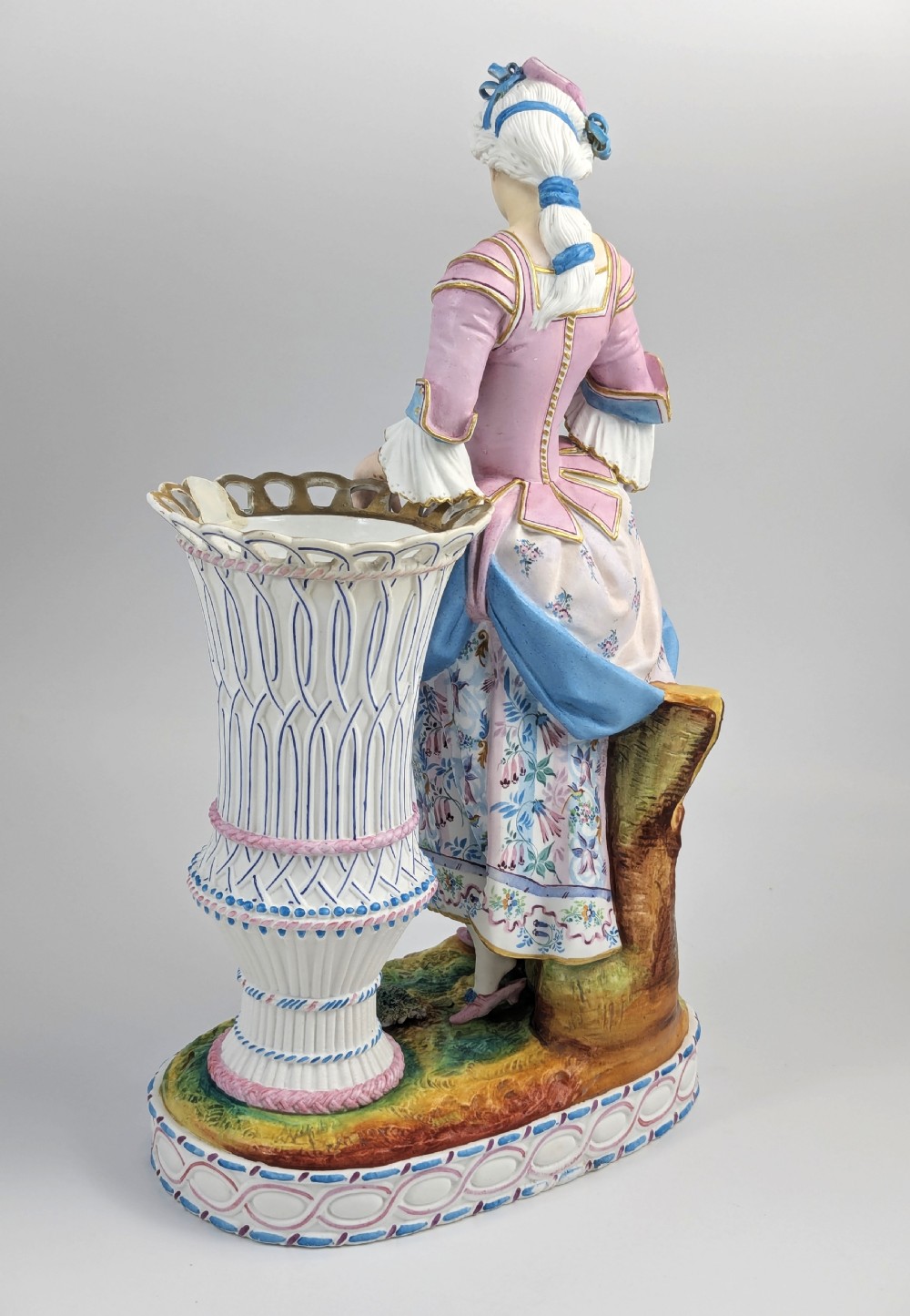 A 19th-century French bisque porcelain figural vase featuring a female figure in pastel Rococo costume standing beside a lattice-patterned vase on a painted base. View IV