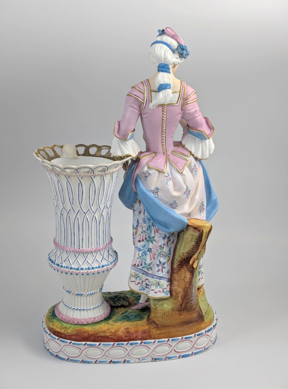 A 19th-century French bisque porcelain figural vase featuring a female figure in pastel Rococo costume standing beside a lattice-patterned vase on a painted base. View V