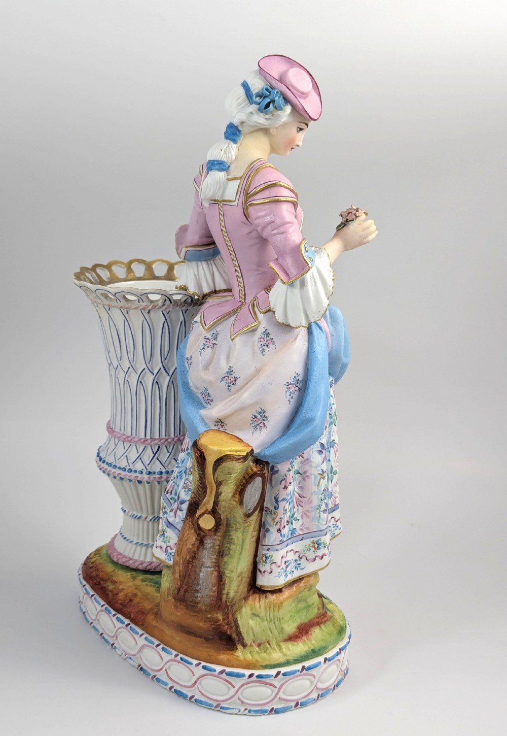 A 19th-century French bisque porcelain figural vase featuring a female figure in pastel Rococo costume standing beside a lattice-patterned vase on a painted base. View VI