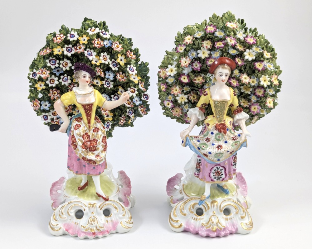 A pair of 19th-century Samson of Paris hand-painted porcelain figures standing against dense, flowering green bocage backgrounds on scrollwork bases.
