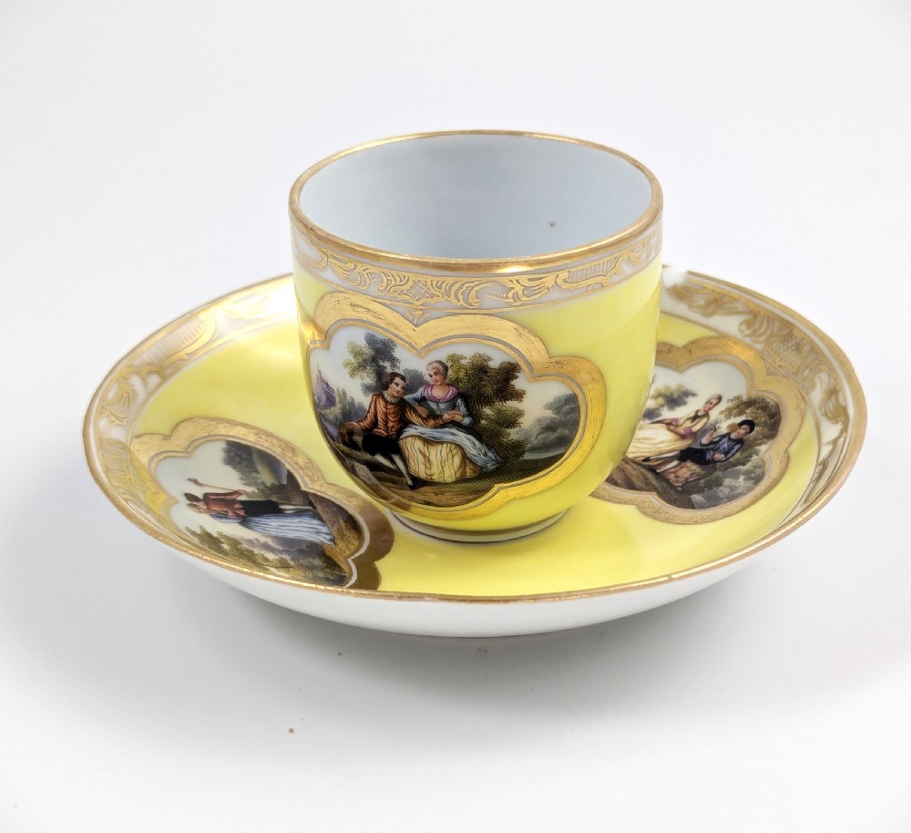 An antique nineteenth-century Meissen porcelain demitasse cup and saucer in yellow featuring hand-painted pastoral scenes set within intricate gilded cartouches. View II