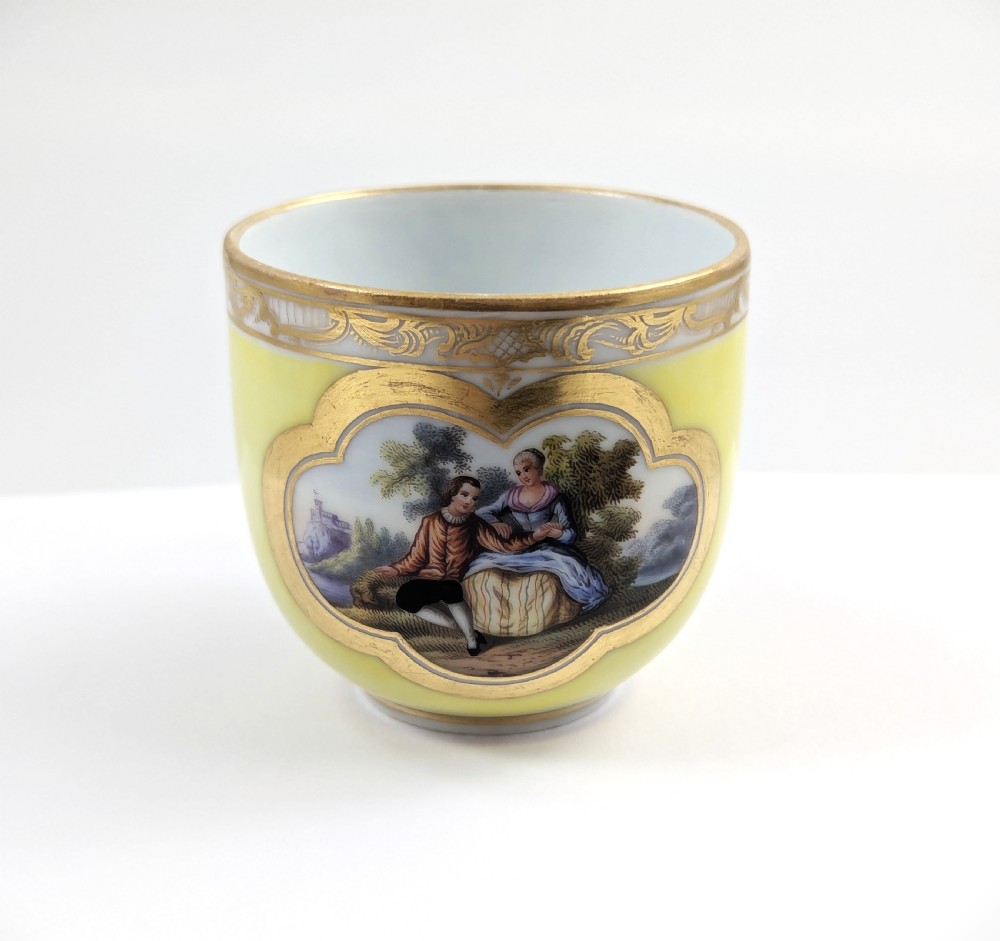 An antique nineteenth-century Meissen porcelain demitasse cup and saucer in yellow featuring hand-painted pastoral scenes set within intricate gilded cartouches. View V