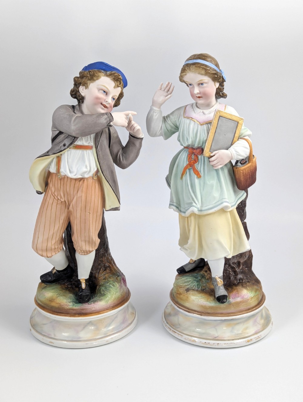 A pair of 19th-century French bisque porcelain figurines depicting a schoolboy and girl in traditional attire on round bases.