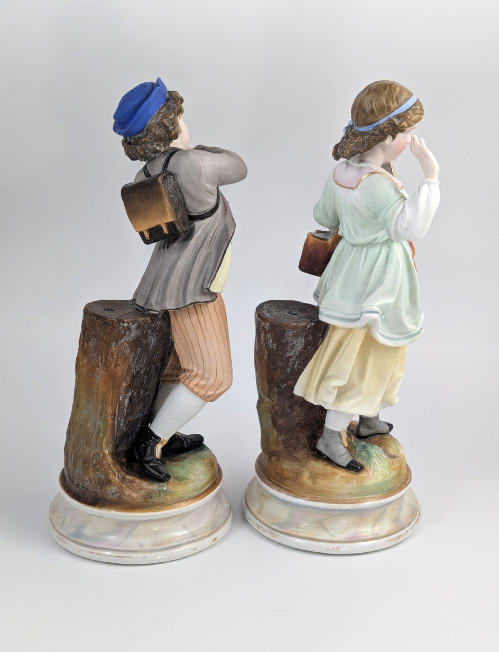 A pair of 19th-century French bisque porcelain figurines depicting a schoolboy and girl in traditional attire on round bases. View IV