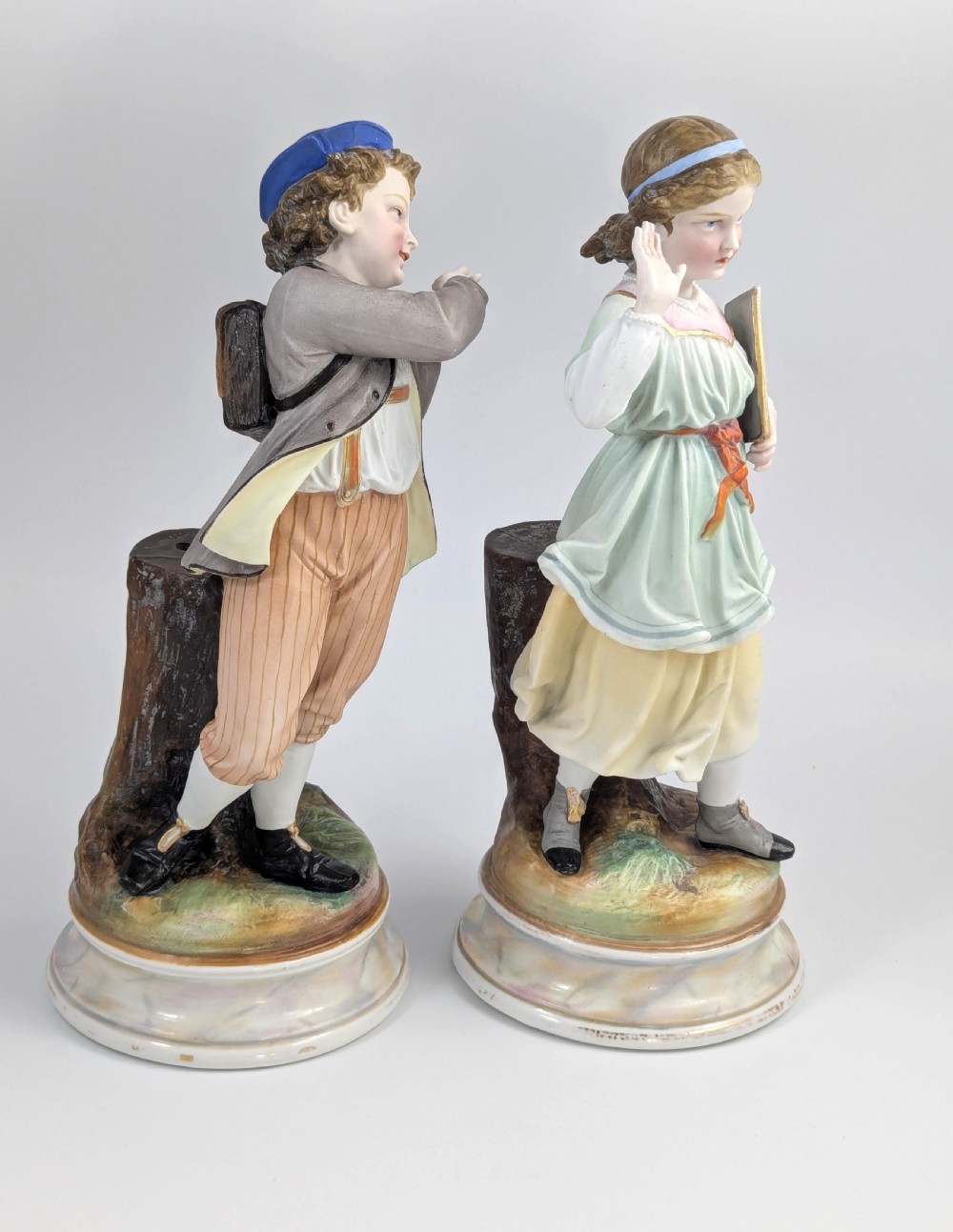 A pair of 19th-century French bisque porcelain figurines depicting a schoolboy and girl in traditional attire on round bases. View V