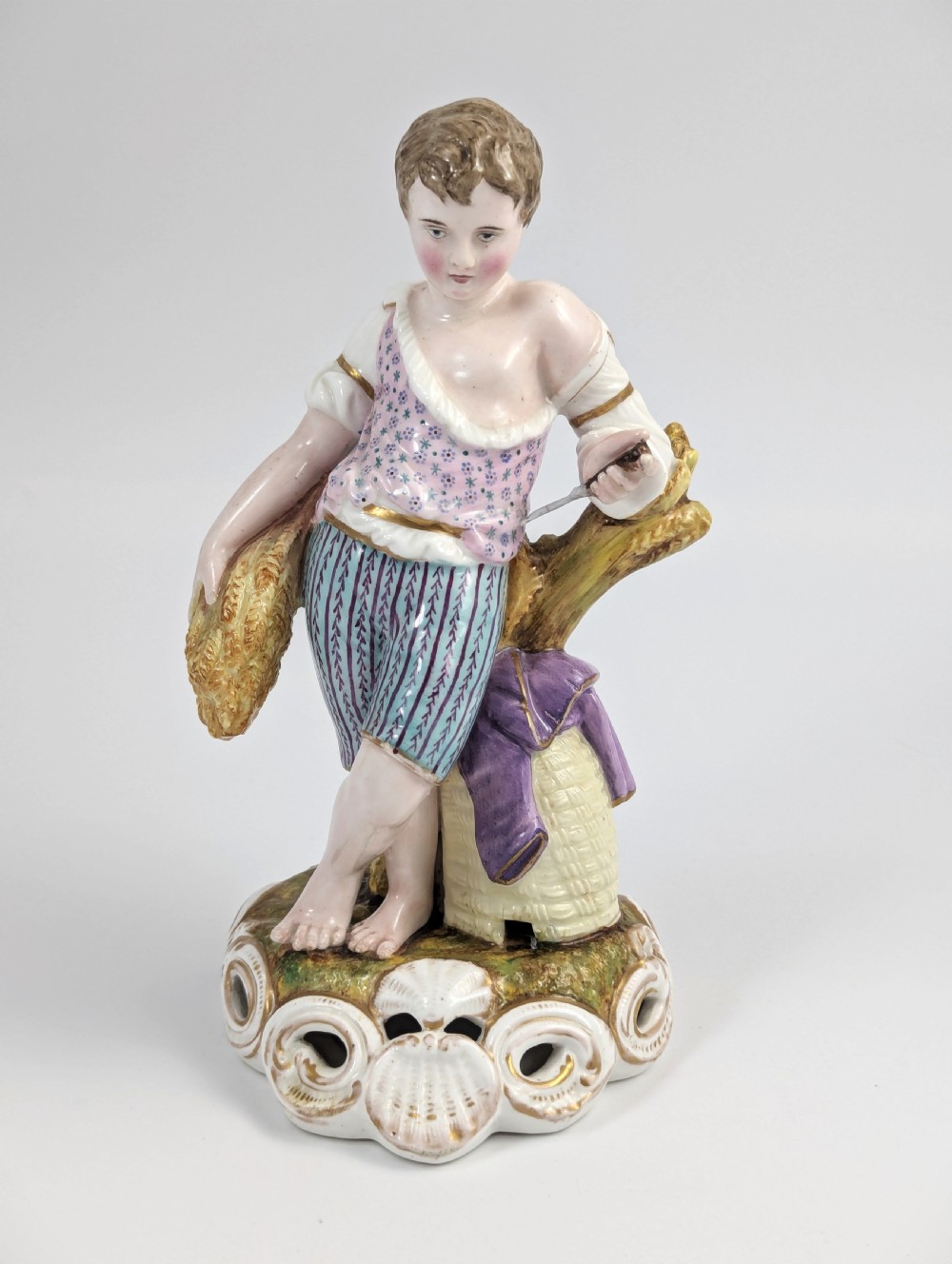 A 19th-century porcelain figurine depicting a young boy holding a sheaf of grain, dressed in a patterned pink top and striped trousers, standing on a scrollwork base with shell motifs.