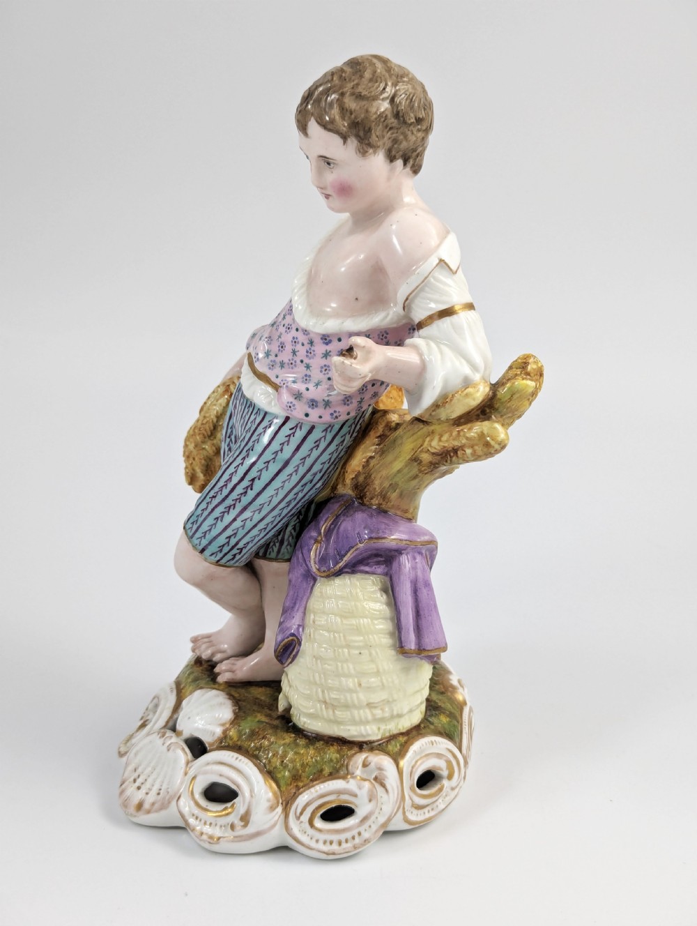 A 19th-century porcelain figurine depicting a young boy holding a sheaf of grain, dressed in a patterned pink top and striped trousers, standing on a scrollwork base with shell motifs. View II