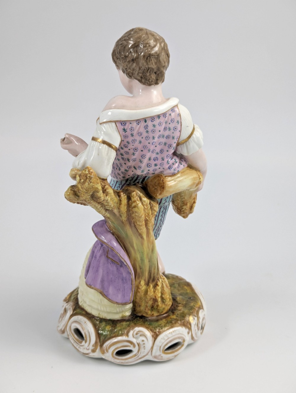 A 19th-century porcelain figurine depicting a young boy holding a sheaf of grain, dressed in a patterned pink top and striped trousers, standing on a scrollwork base with shell motifs. View III