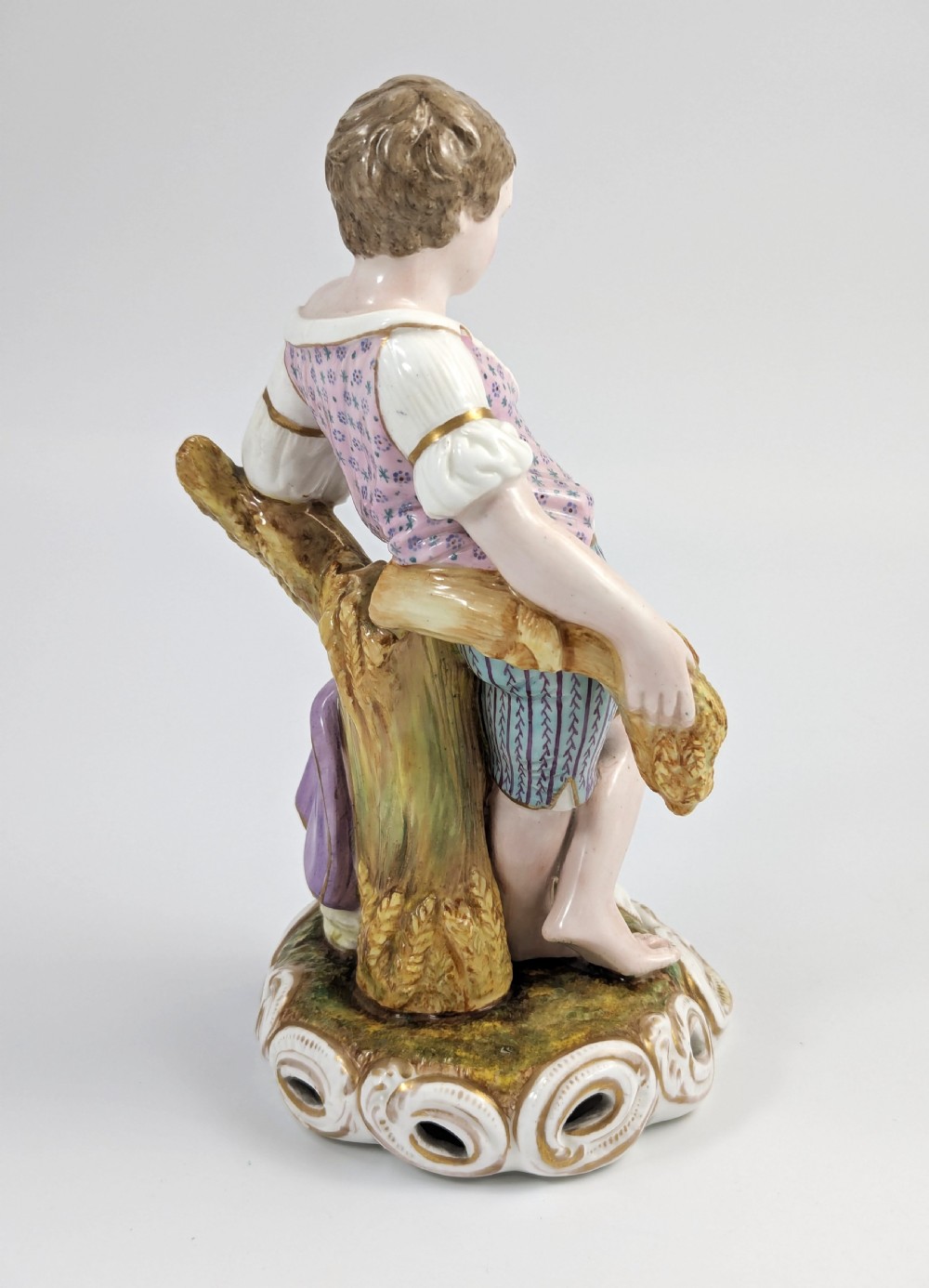 A 19th-century porcelain figurine depicting a young boy holding a sheaf of grain, dressed in a patterned pink top and striped trousers, standing on a scrollwork base with shell motifs. View IV