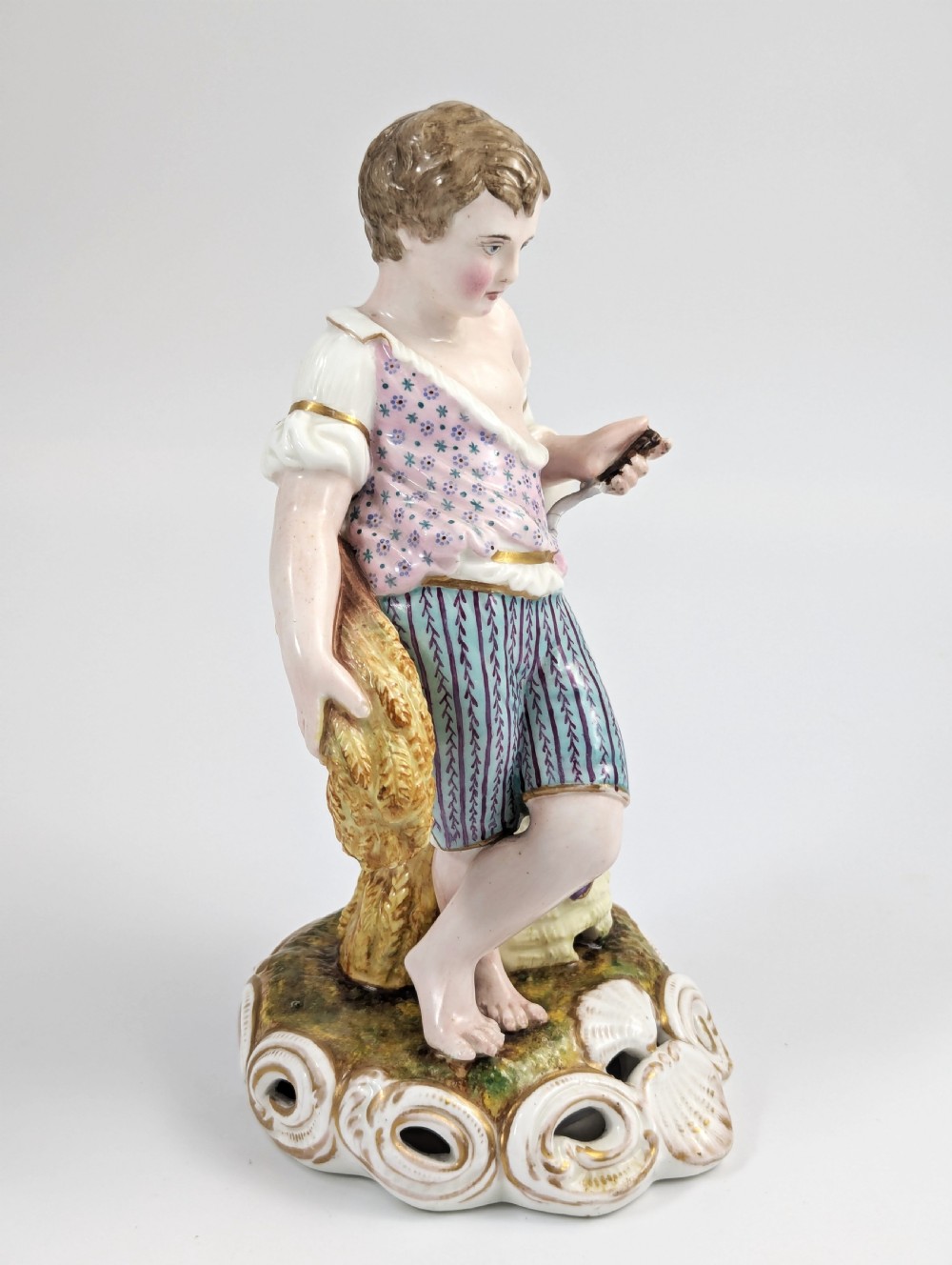 A 19th-century porcelain figurine depicting a young boy holding a sheaf of grain, dressed in a patterned pink top and striped trousers, standing on a scrollwork base with shell motifs. View V