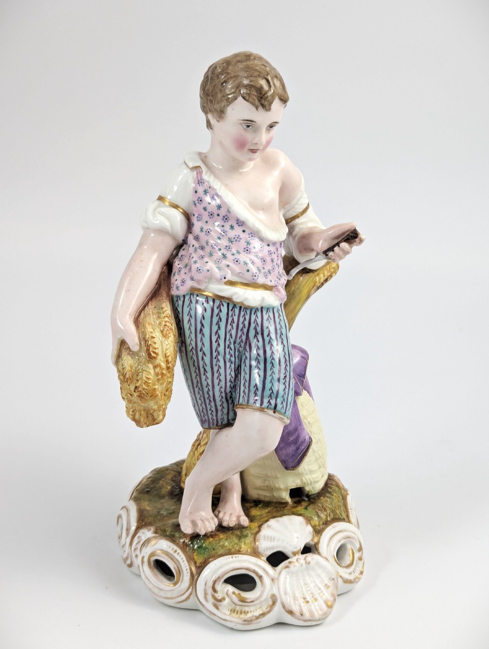 A 19th-century porcelain figurine depicting a young boy holding a sheaf of grain, dressed in a patterned pink top and striped trousers, standing on a scrollwork base with shell motifs. View VI