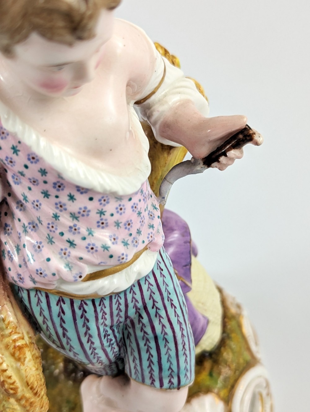 A 19th-century porcelain figurine depicting a young boy holding a sheaf of grain, dressed in a patterned pink top and striped trousers, standing on a scrollwork base with shell motifs. View VII