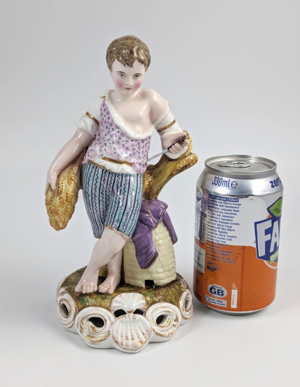 A 19th-century porcelain figurine depicting a young boy holding a sheaf of grain, dressed in a patterned pink top and striped trousers, standing on a scrollwork base with shell motifs. View IX