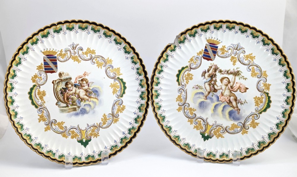 A pair of 18th-century French tin-glazed earthenware display plates with hand-painted putti motifs, armorial shields, and green scalloped borders.
