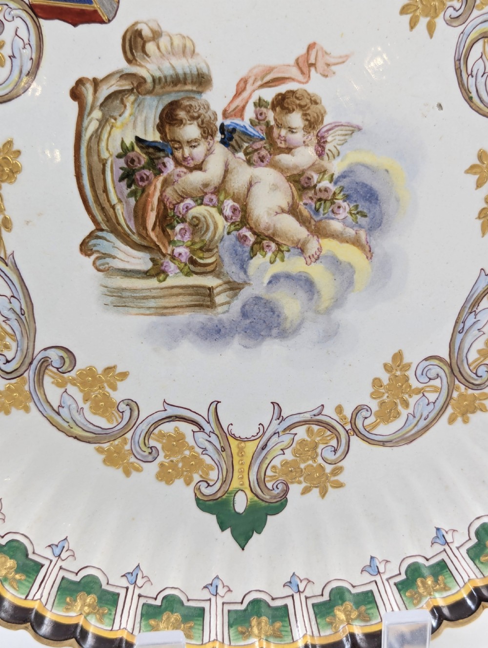 A pair of 18th-century French tin-glazed earthenware display plates with hand-painted putti motifs, armorial shields, and green scalloped borders. View VIII