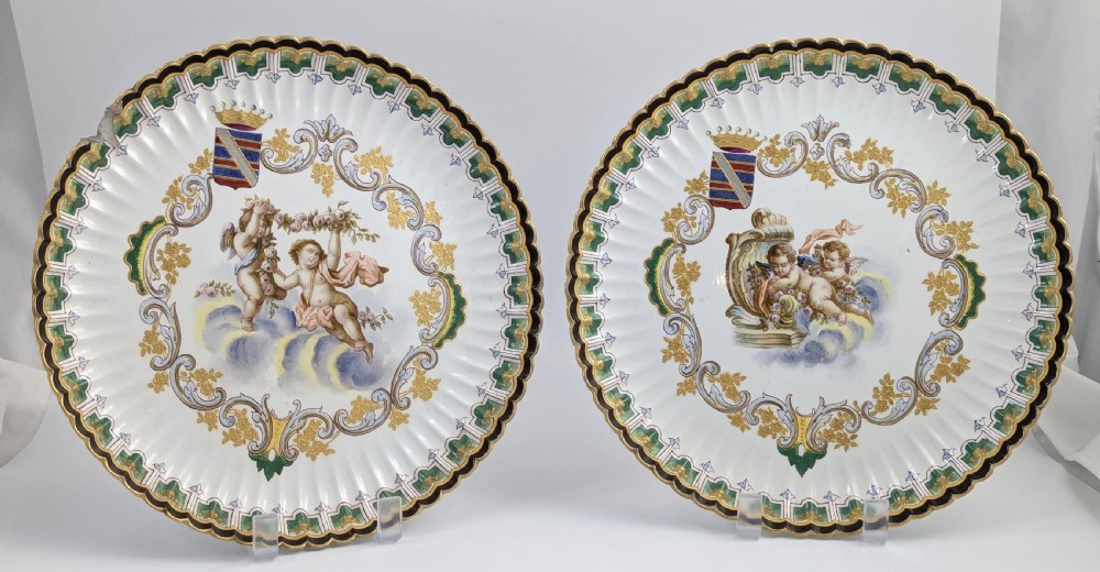 A pair of 18th-century French tin-glazed earthenware display plates with hand-painted putti motifs, armorial shields, and green scalloped borders. View X