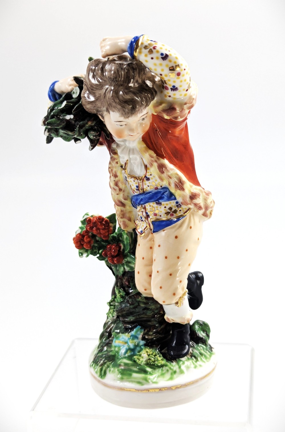 An 18th-century Derby porcelain figurine representing Winter, featuring a person in a red coat and fur-lined waistcoat carrying firewood on a green foliage base.