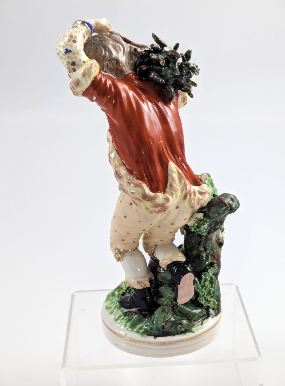 An 18th-century Derby porcelain figurine representing Winter, featuring a person in a red coat and fur-lined waistcoat carrying firewood on a green foliage base. View IV