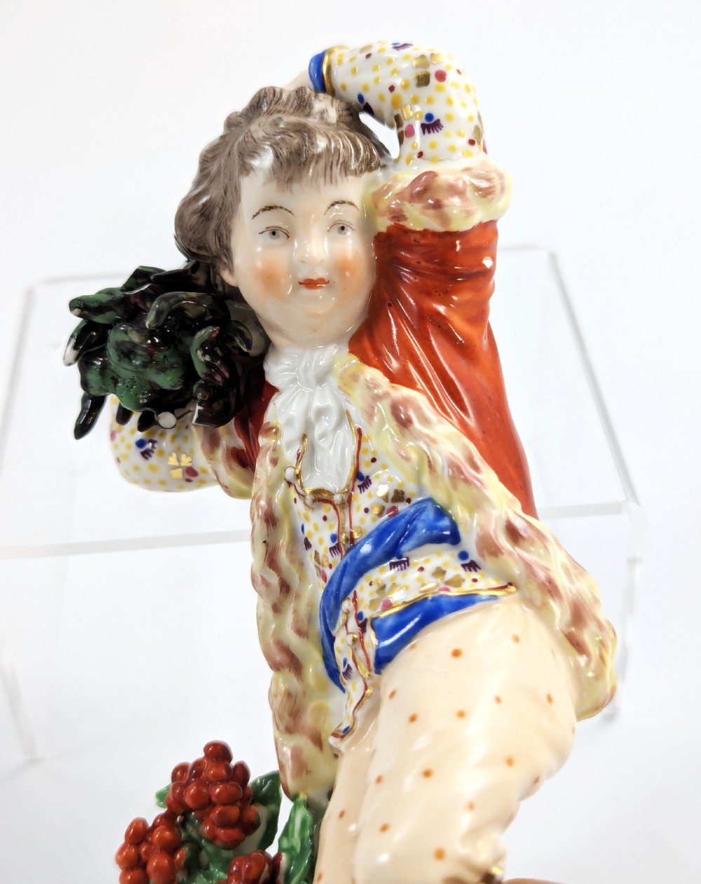 An 18th-century Derby porcelain figurine representing Winter, featuring a person in a red coat and fur-lined waistcoat carrying firewood on a green foliage base. View VIII