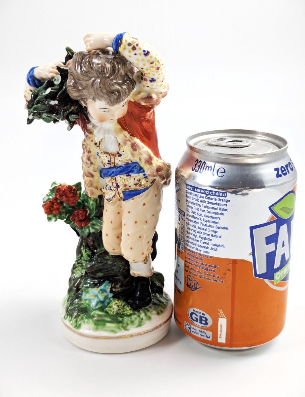 An 18th-century Derby porcelain figurine representing Winter, featuring a person in a red coat and fur-lined waistcoat carrying firewood on a green foliage base. View X