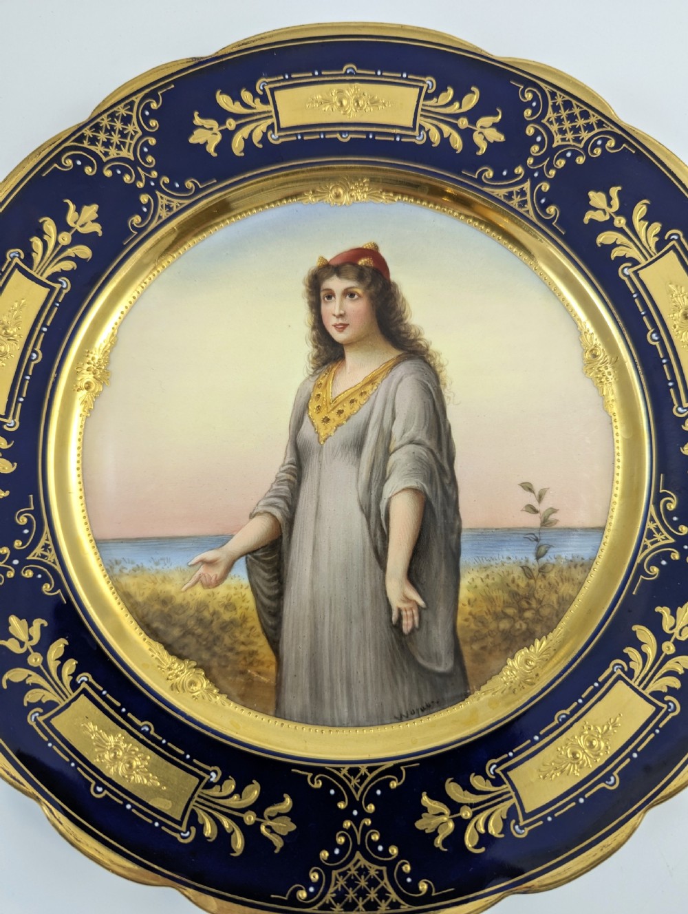 A circular porcelain cabinet plate with a central hand-painted portrait of a woman in draped attire set within a cobalt blue rim embellished with gold gilding. View IX