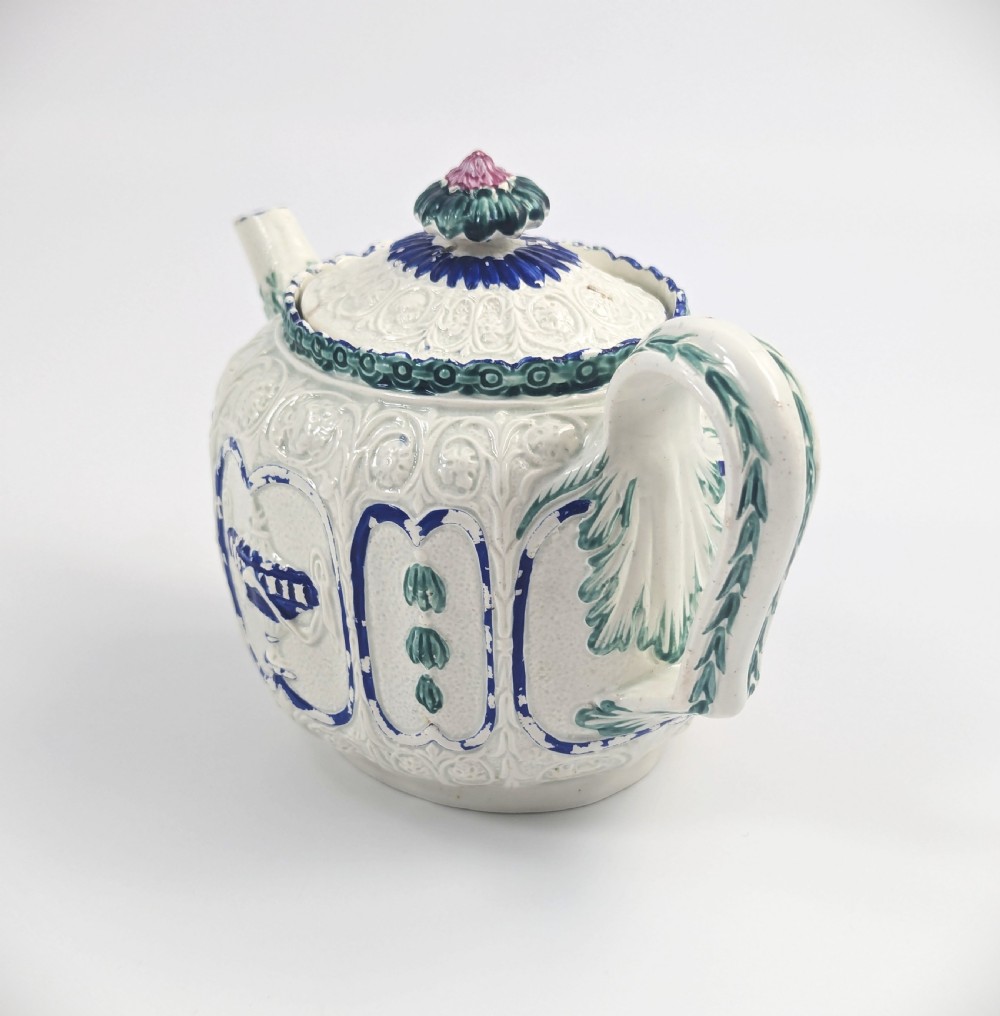 An 18th-century lead-glazed ceramic teapot featuring relief-moulded arches, blue and green painted accents, and a moulded lid with a pink flower finial. View II